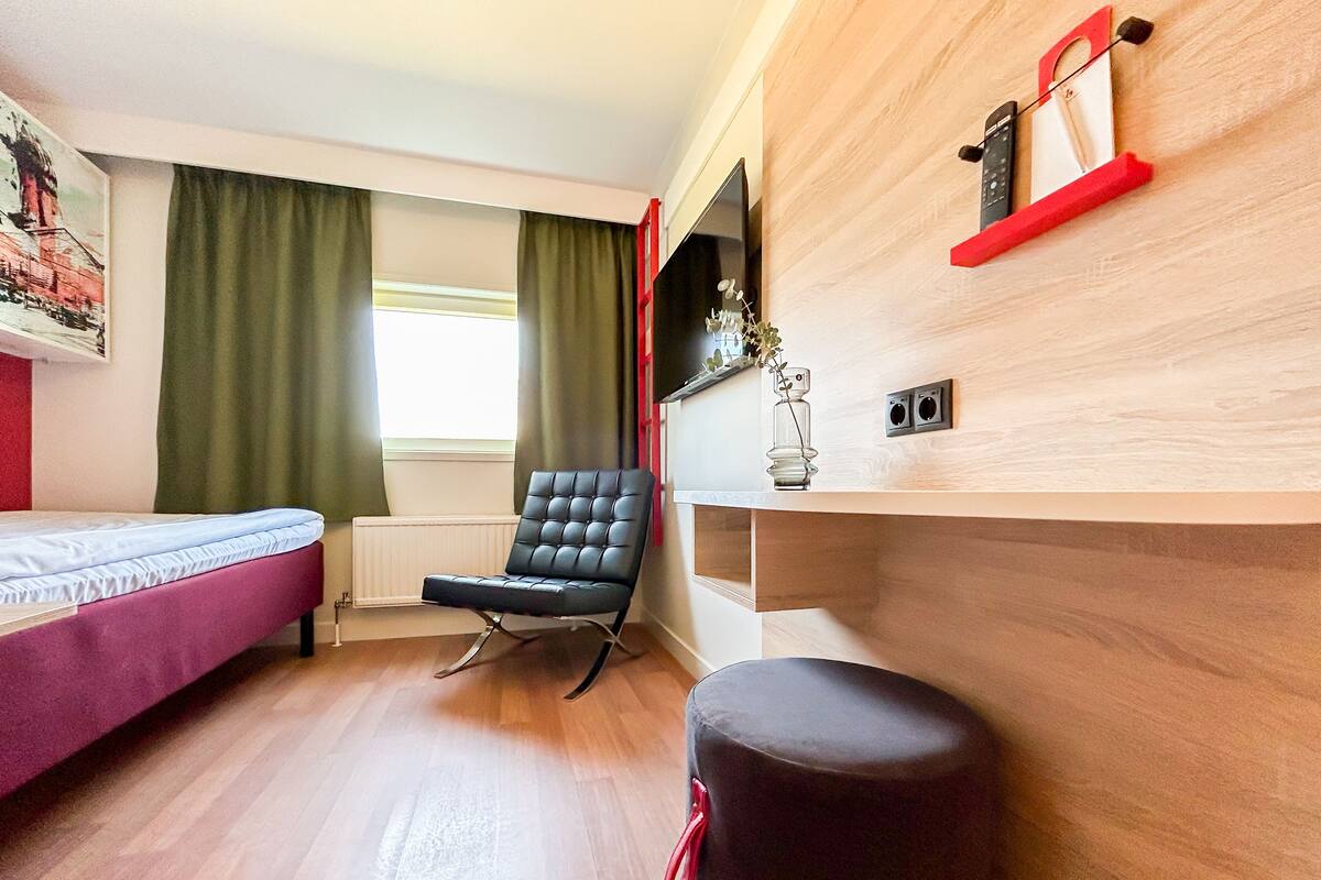 A modern room with a bed, a black chair, a small stool, a radiator, and a shelf with a red object.