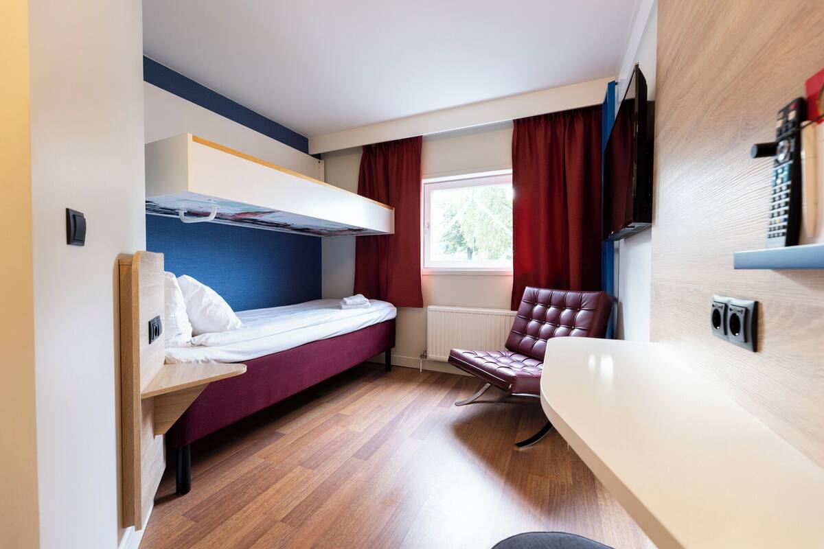 A compact hotel room with a bunk bed, a single bed, a leather armchair, a TV, and a window with curtains.