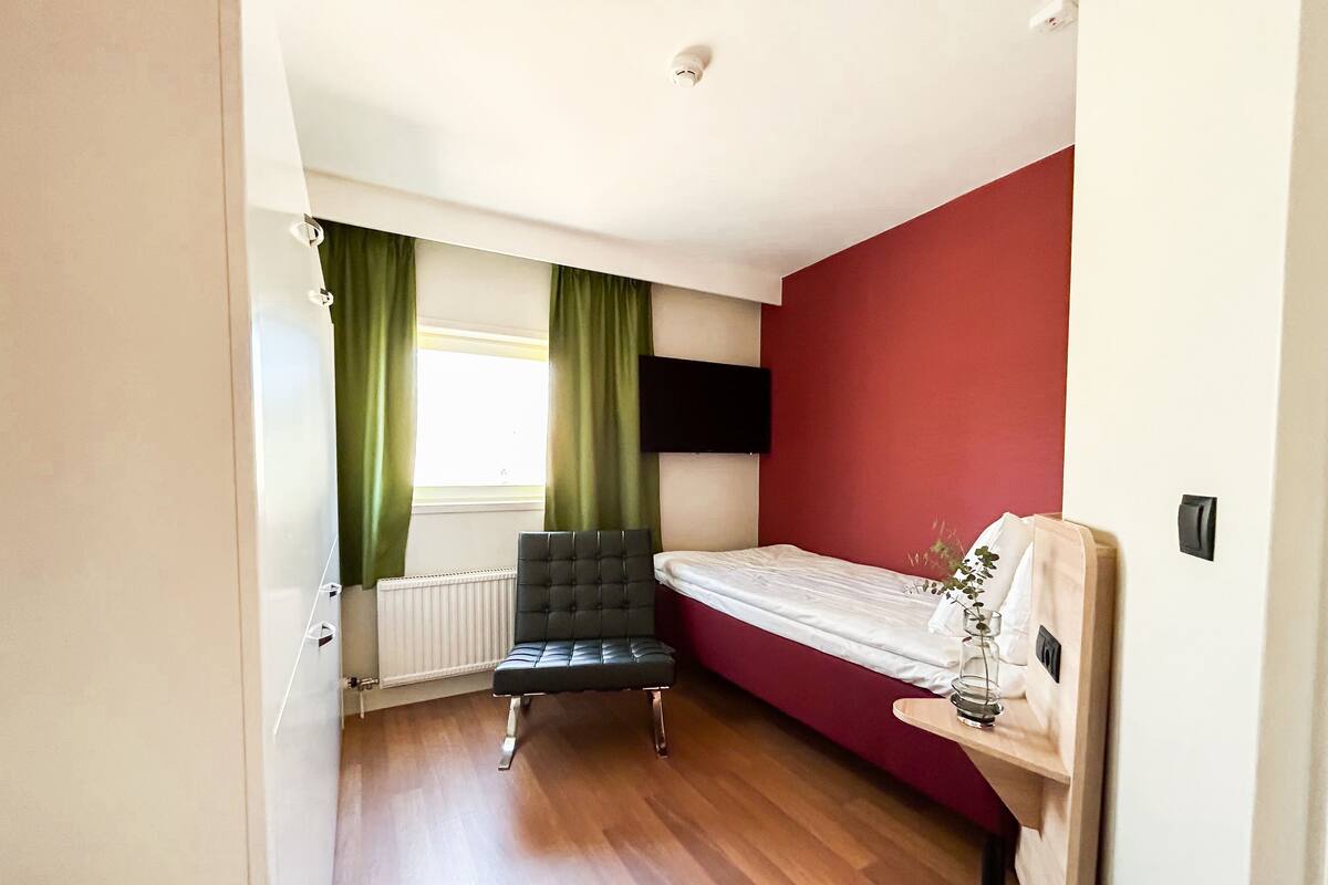 A bedroom with a bed, a chair, a radiator, a window with curtains, and a wall-mounted TV.