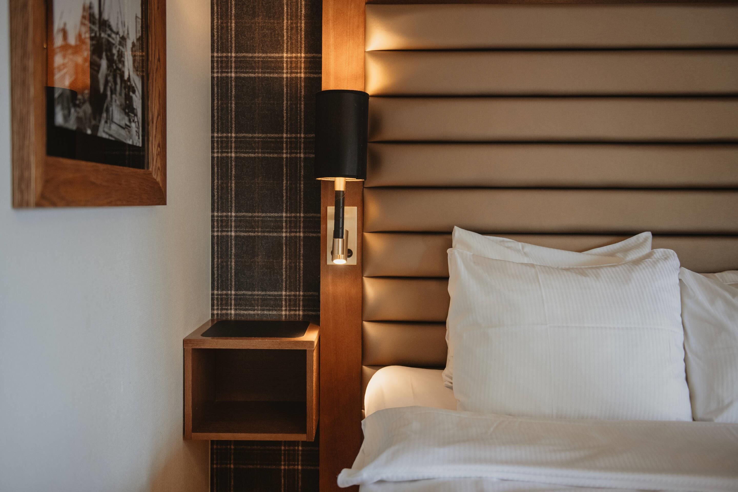 A neatly made bed with white linens, a wooden nightstand with a black lamp, and a plaid-patterned wall hanging.