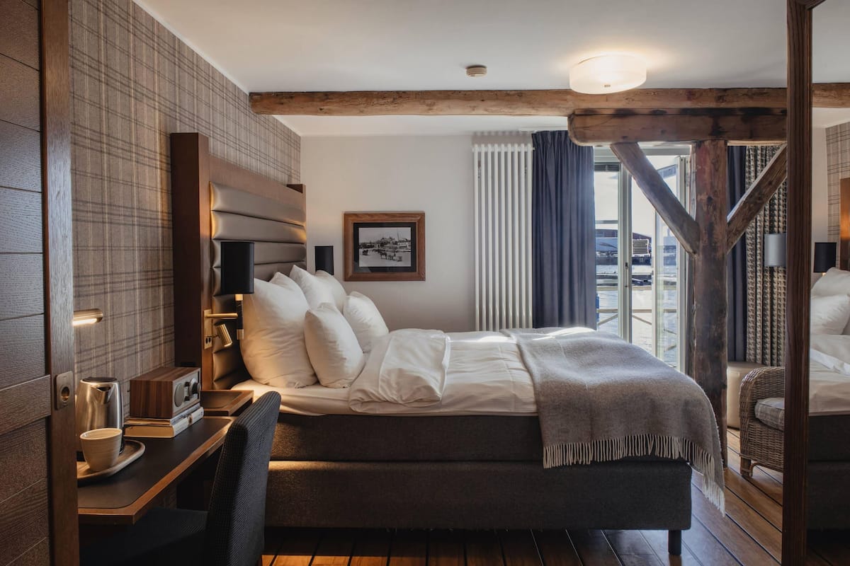 A modern hotel room with a large bed, a wooden headboard, a small desk with a chair, and a window with curtains.