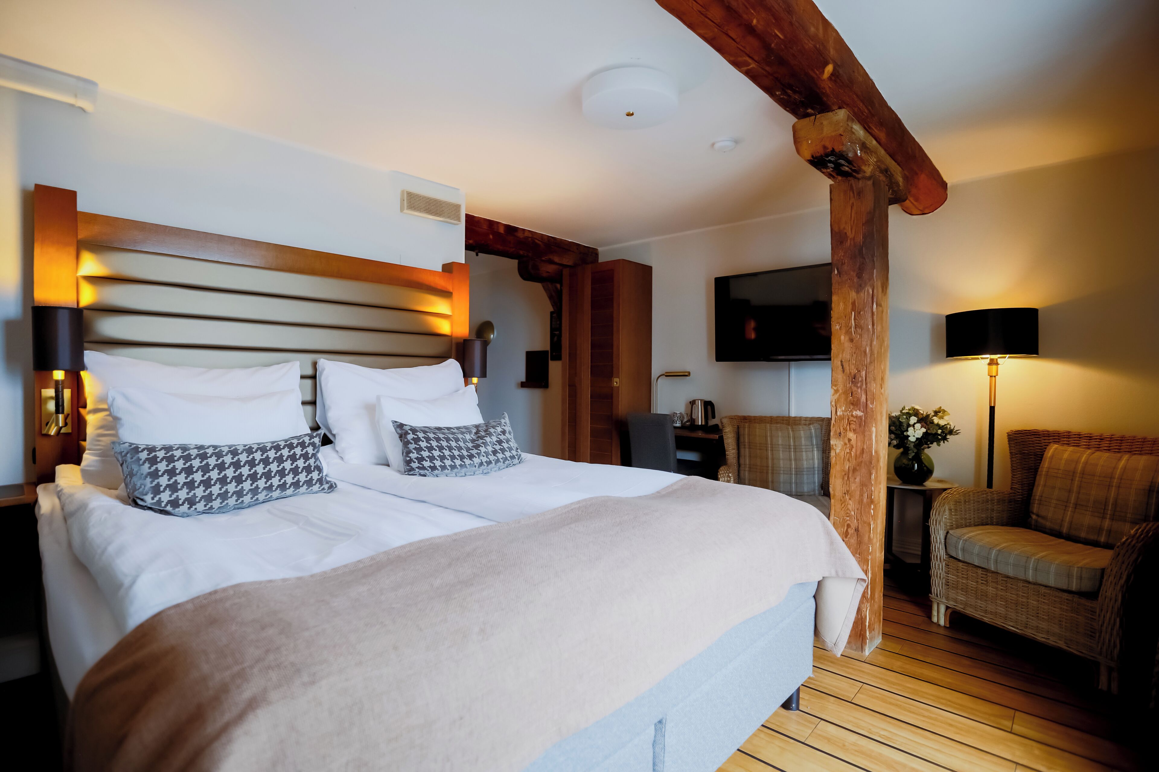 Quarto com cama grande, vigas de madeira, televisão, cadeira e mesa de cabeceira.