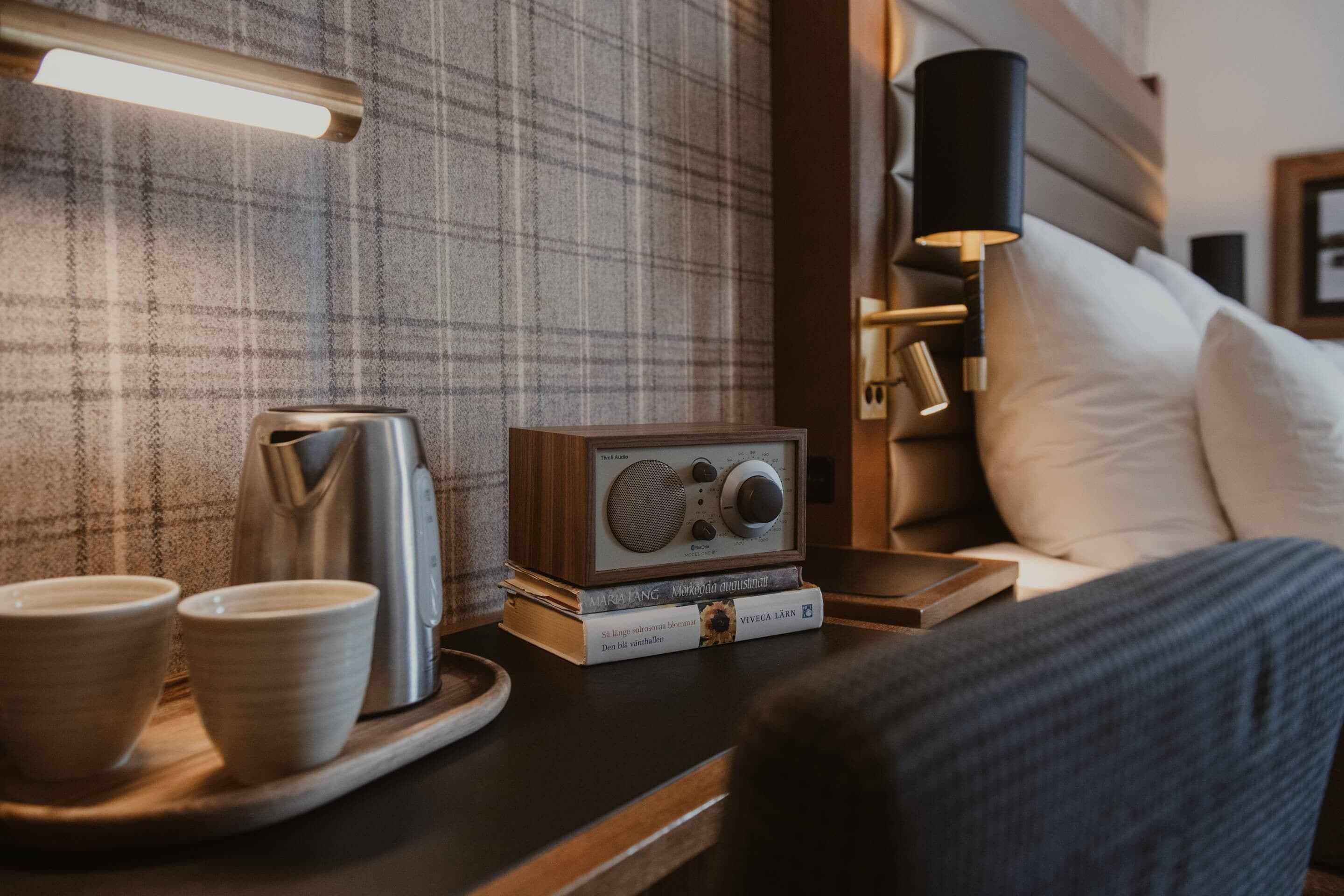 A neatly arranged hotel room with a plaid-patterned wall, a bedside table with a radio, a kettle, and two cups.