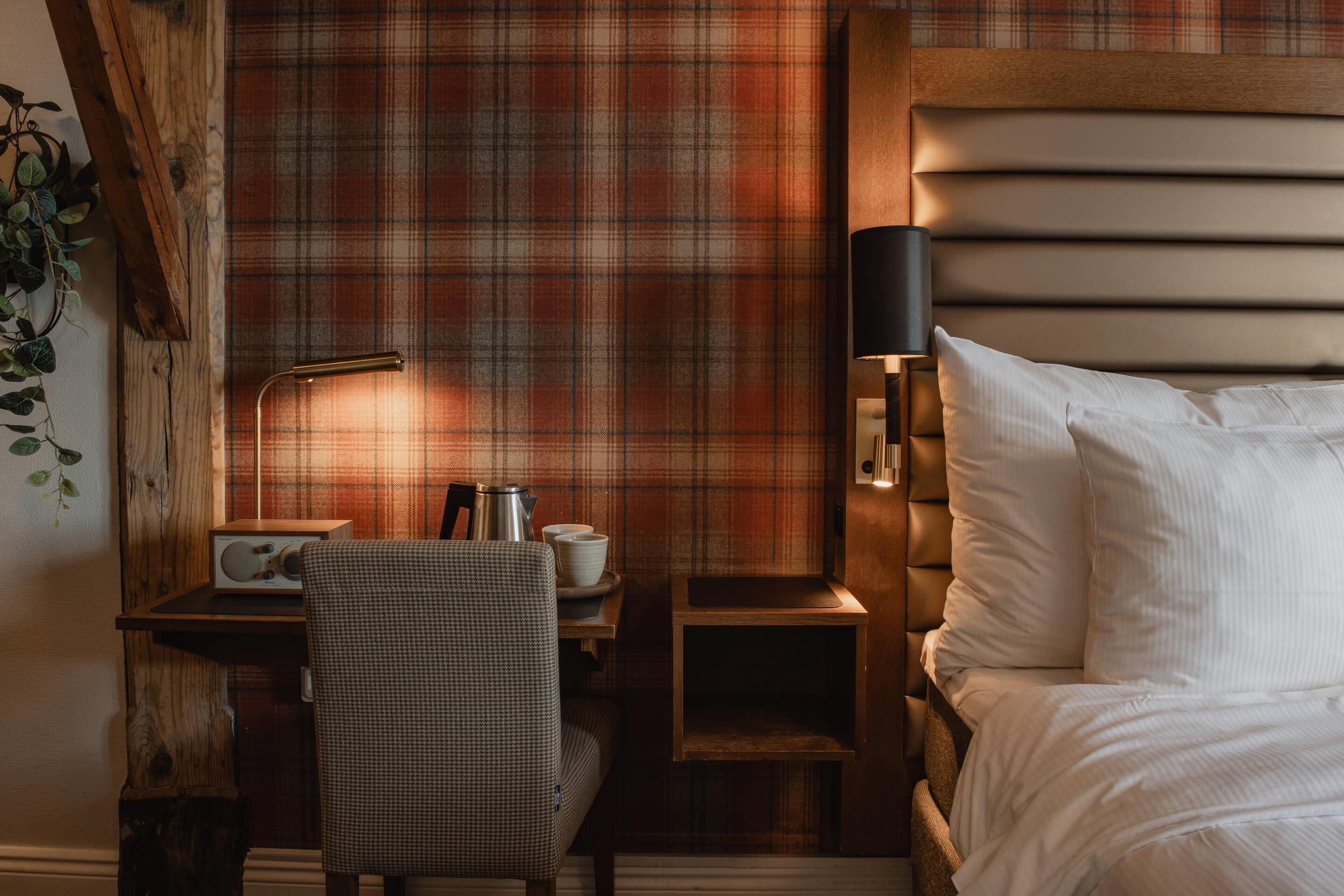 A cozy hotel room with a plaid-patterned wall, a wooden desk with a radio, a chair, a bedside table with a lamp, and a bed with white linens.