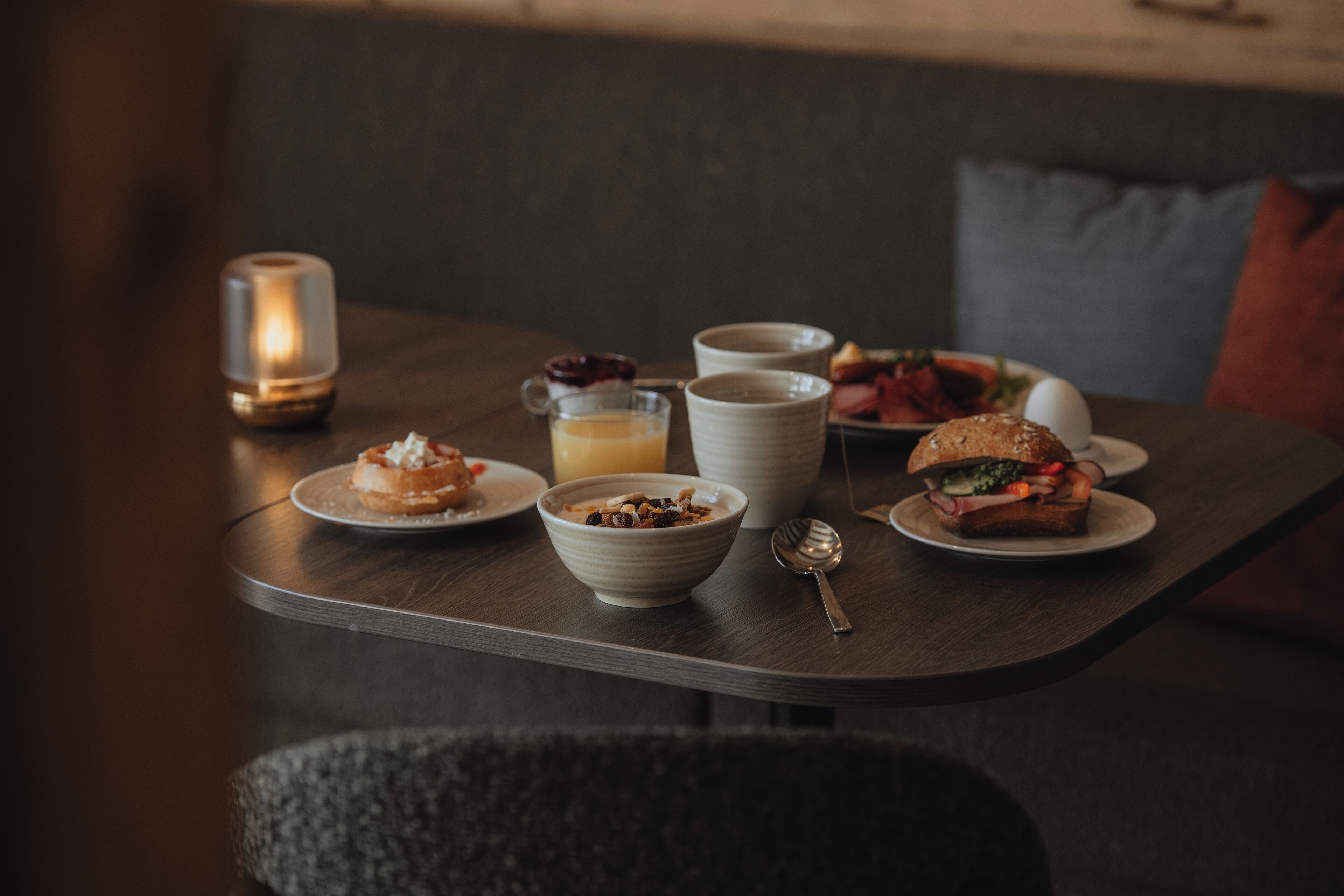 Ein Esstisch mit Kerze, einer Schüssel mit Müsli, einer Kaffeetasse, einem Sandwich und einem Teller mit Obst.