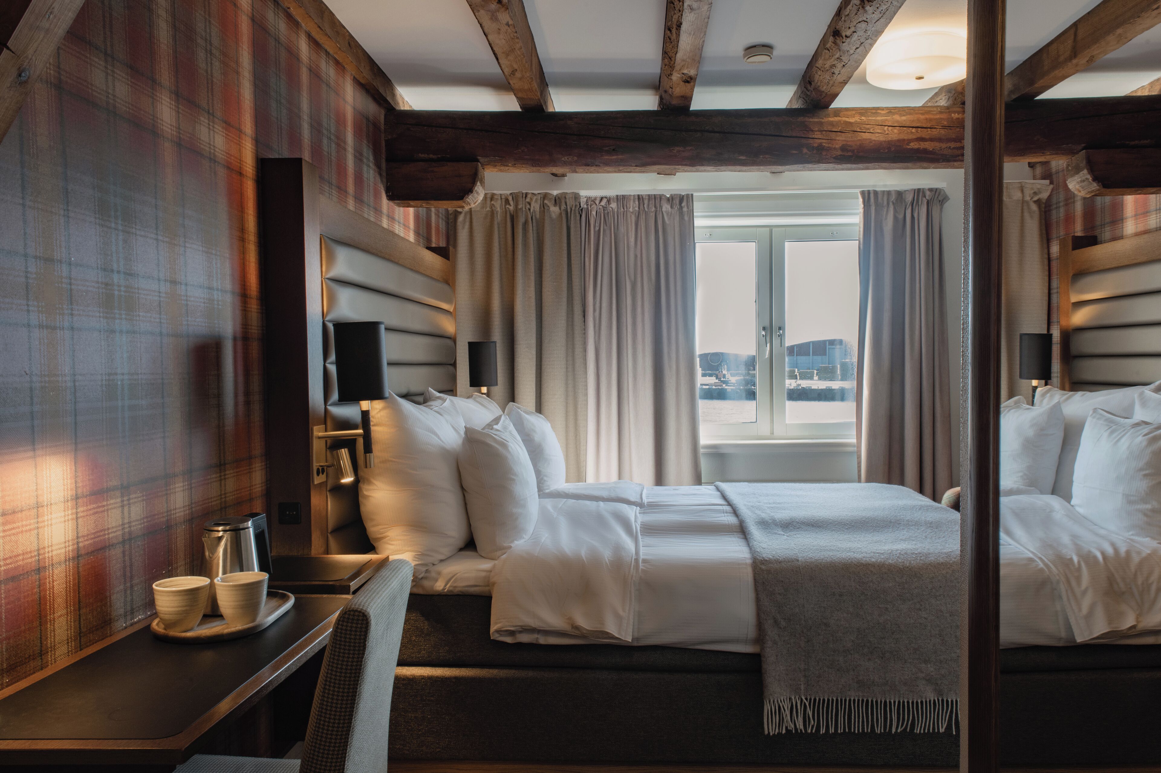A hotel room with a bed, a desk, a chair, a window with curtains, and a view of the city.