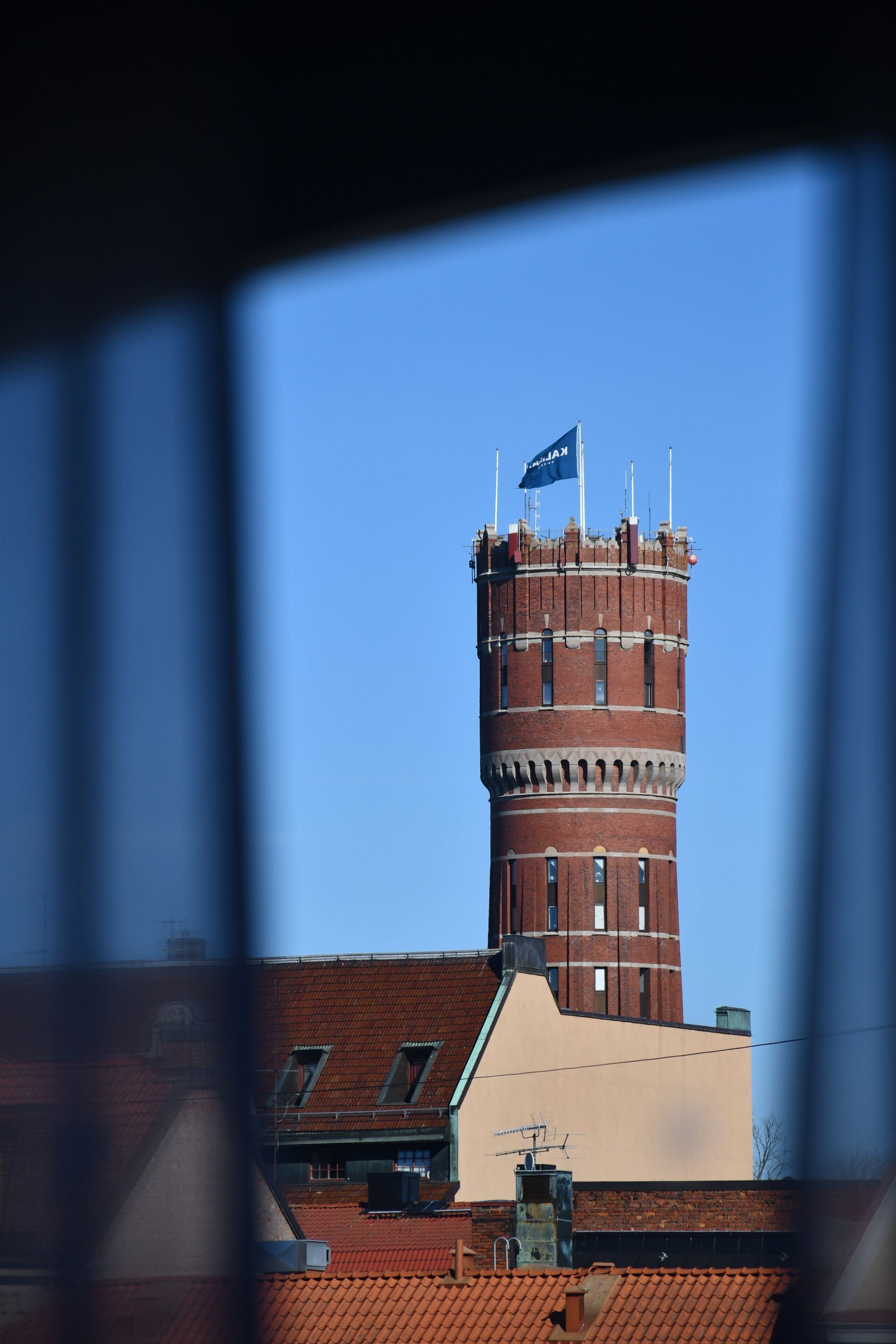 Una torre de ladrillo con una bandera, un edificio con techo rojo y una ventana con vista al cielo.