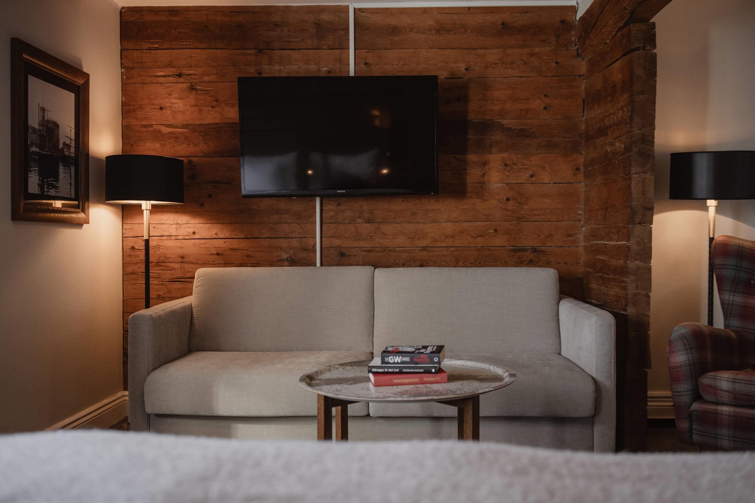 A modern living room with a sofa, a coffee table with books, a TV mounted on a wooden wall, and two floor lamps.