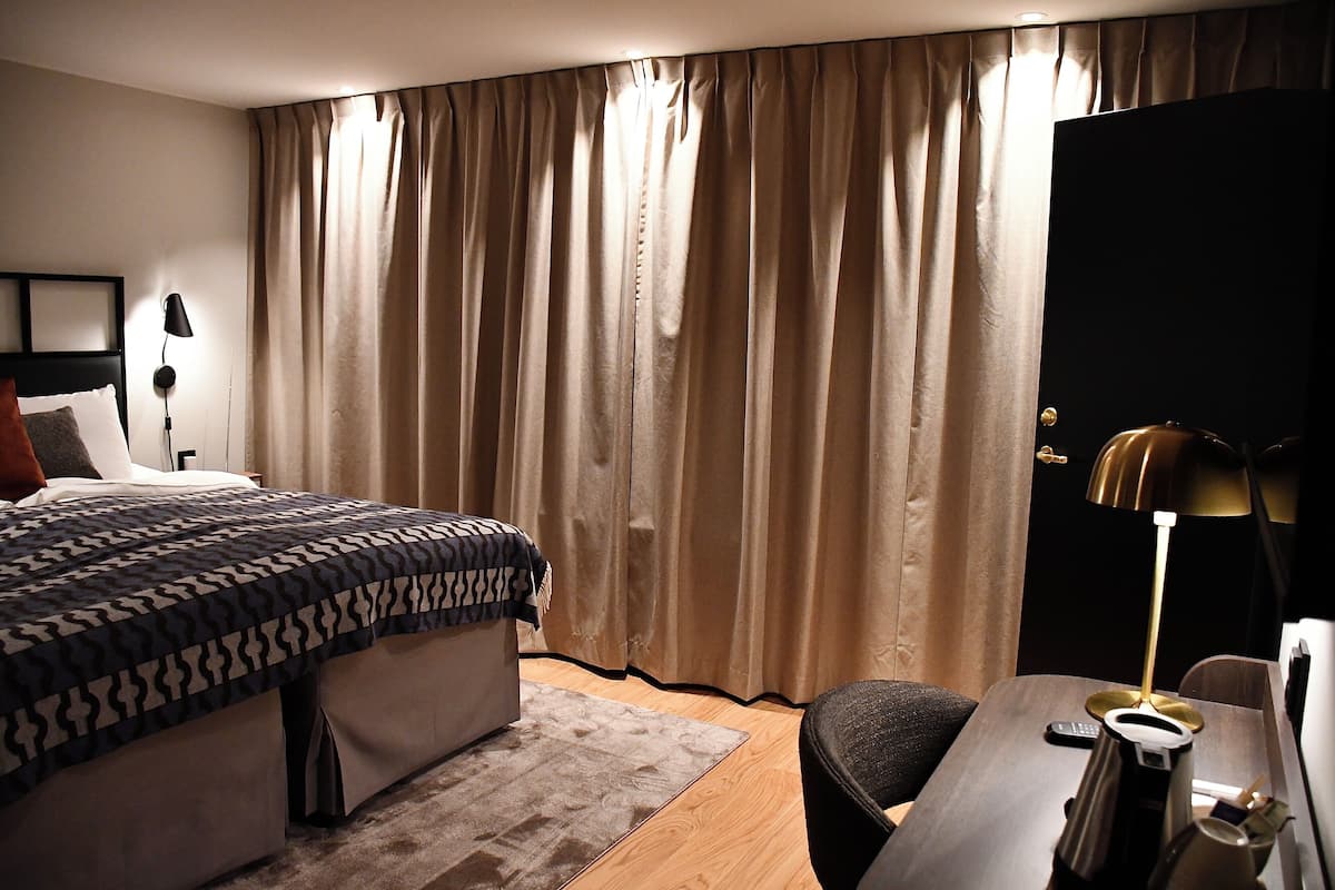 A hotel room with a bed, a desk, a chair, a lamp, and curtains.