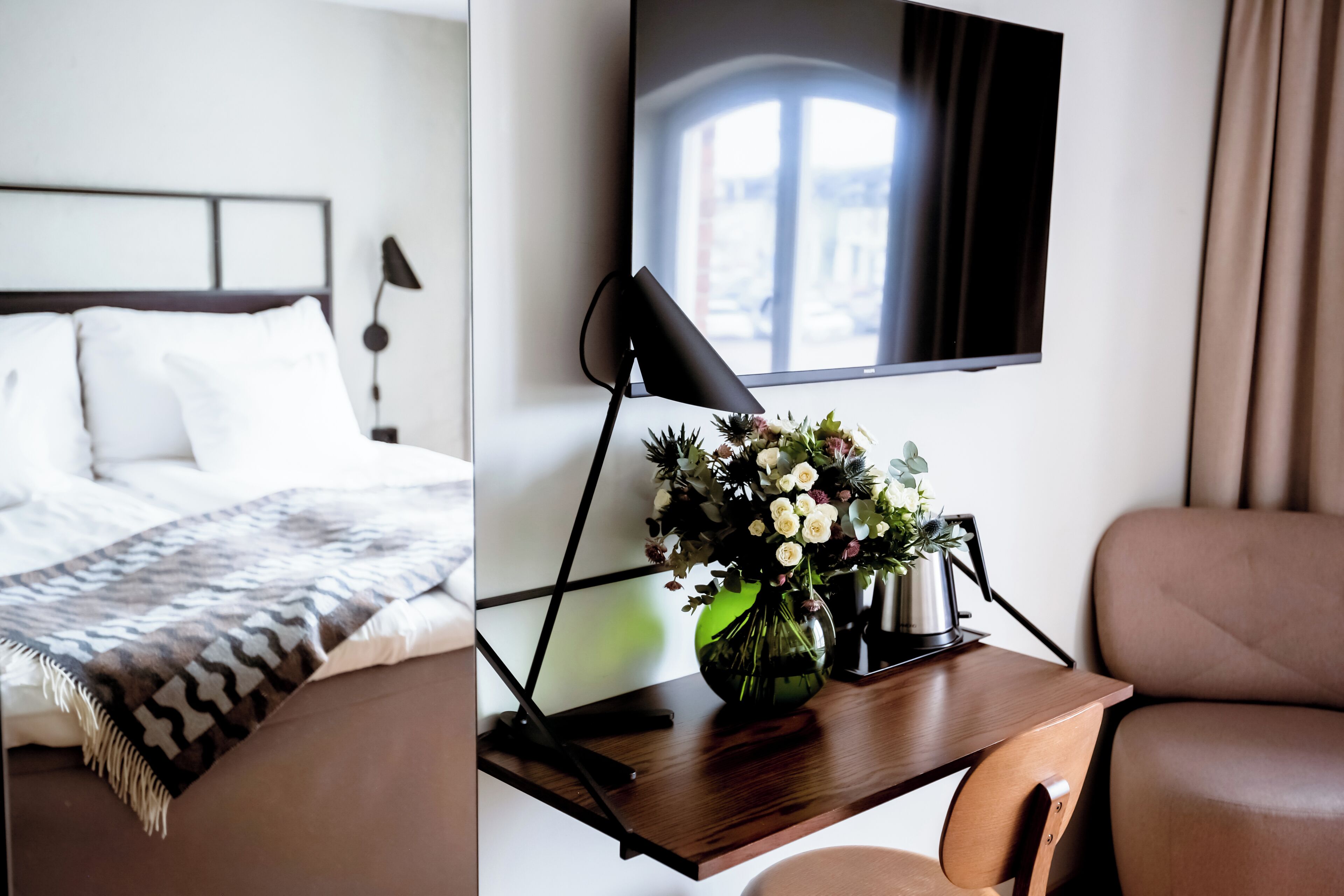 A hotel room with a bed, a desk with a lamp, a vase with flowers, and a TV mounted on the wall.