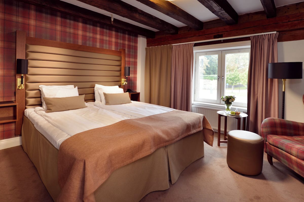 A hotel room with a large bed, a wooden headboard, a small table with a vase, a plaid armchair, and a window with curtains.