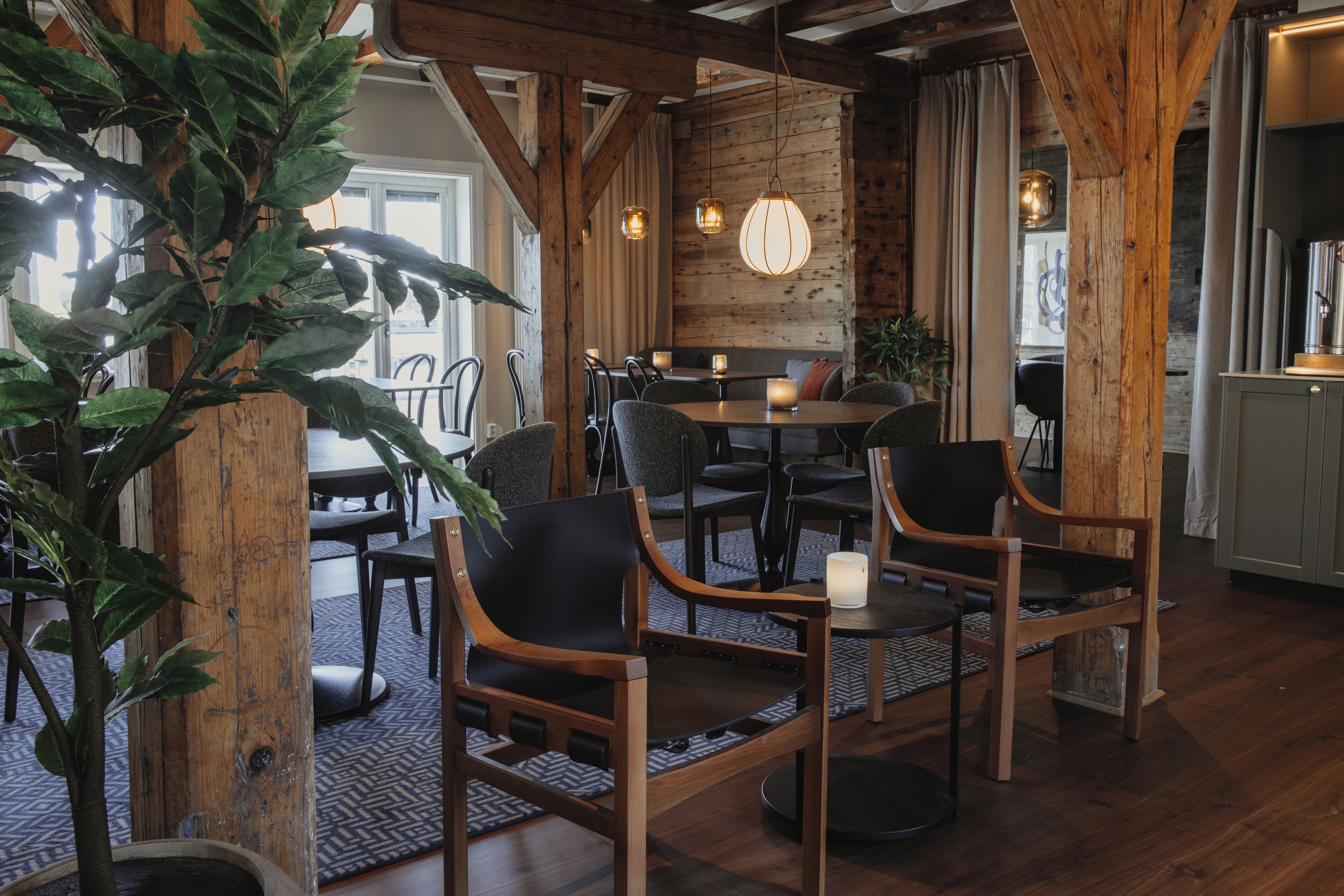 A rustic interior with wooden beams, a dining area with tables and chairs, and a large potted plant.