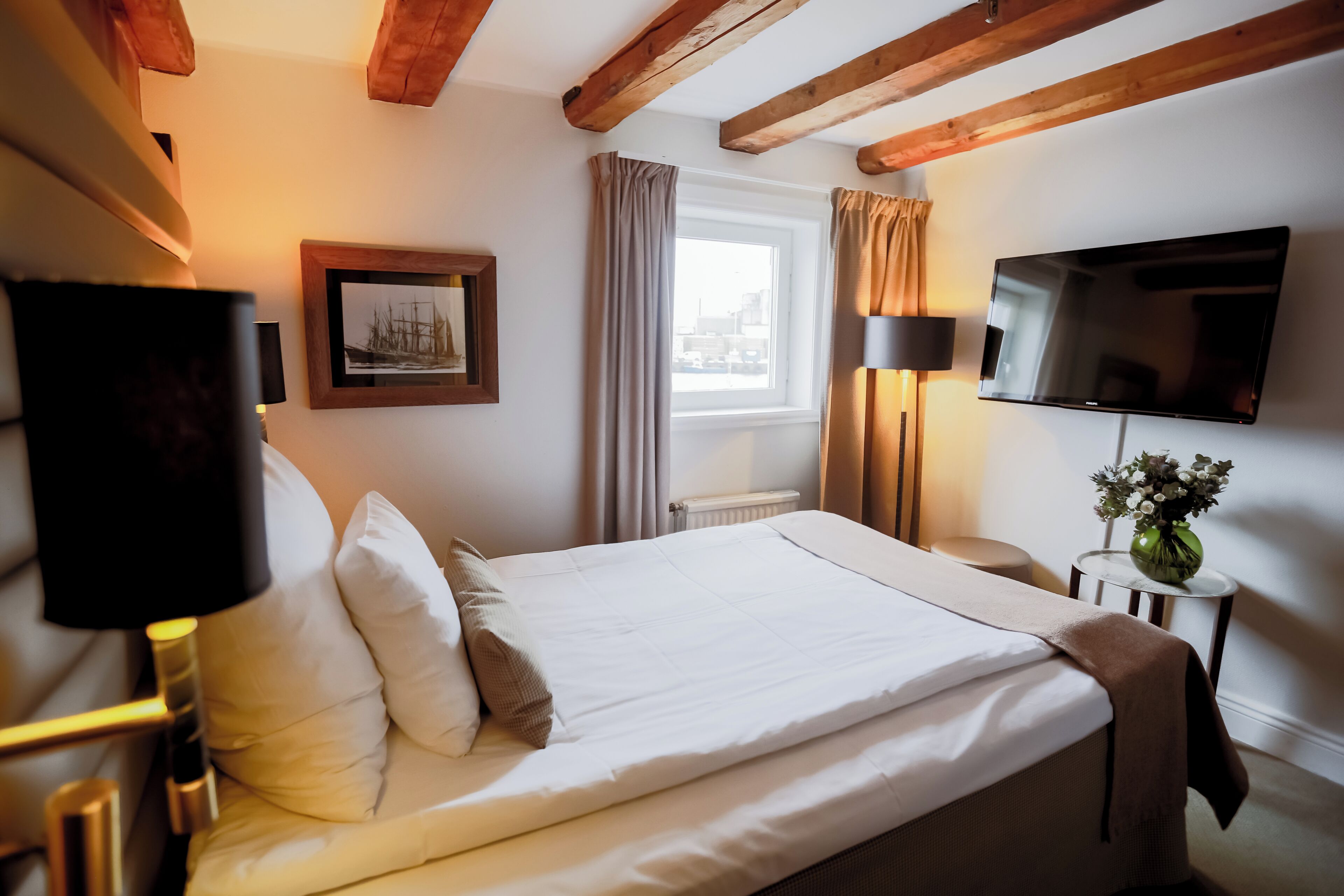 A hotel room with a bed, a TV, a window with curtains, a framed picture, and a small table with a vase.