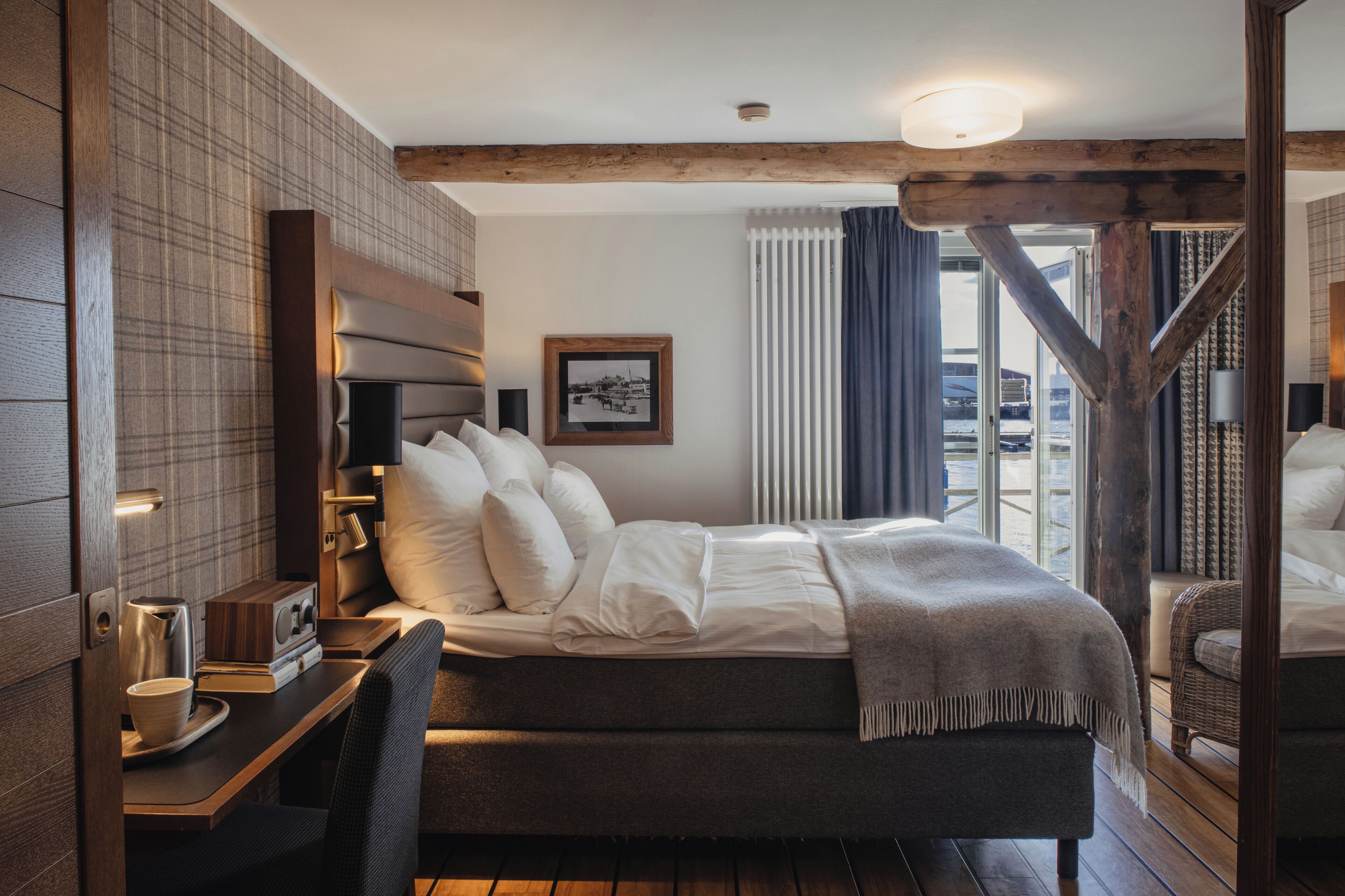 A modern hotel room with a large bed, a wooden headboard, a desk with a chair, and a view of the outside.