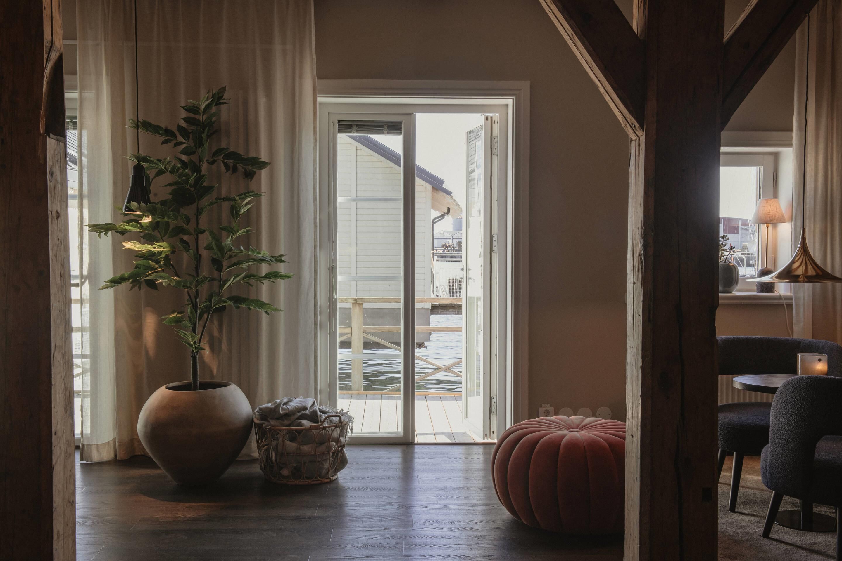 Habitación con una planta grande en maceta, una cesta de mimbre, un otoman redondo y vistas a una terraza exterior a través de puertas corredizas de vidrio.