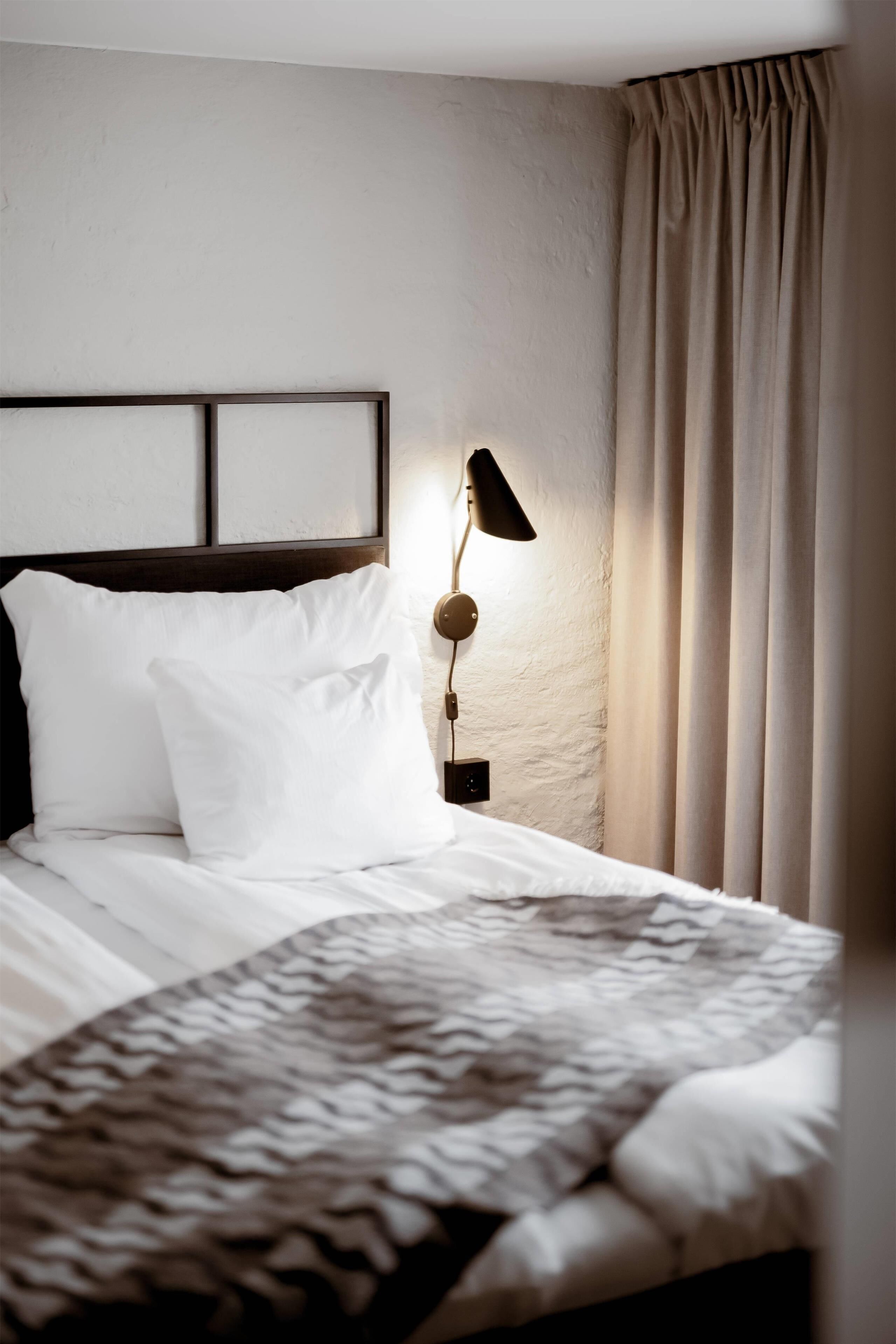 Un lit bien fait, avec du linge de lit blanc et un couvre-lit à motifs, une lampe de chevet et une tête de lit adossée à un mur à texture.
