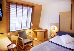 Habitación de hotel con cama, escritorio, silla, mesita y ventana con persianas.