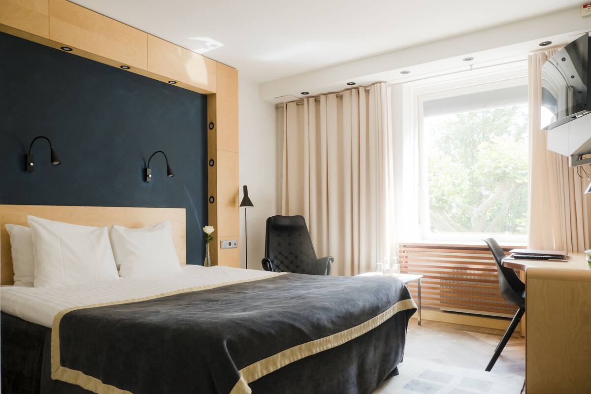 A hotel room with a large bed, a desk, a chair, a TV, and a window with curtains.