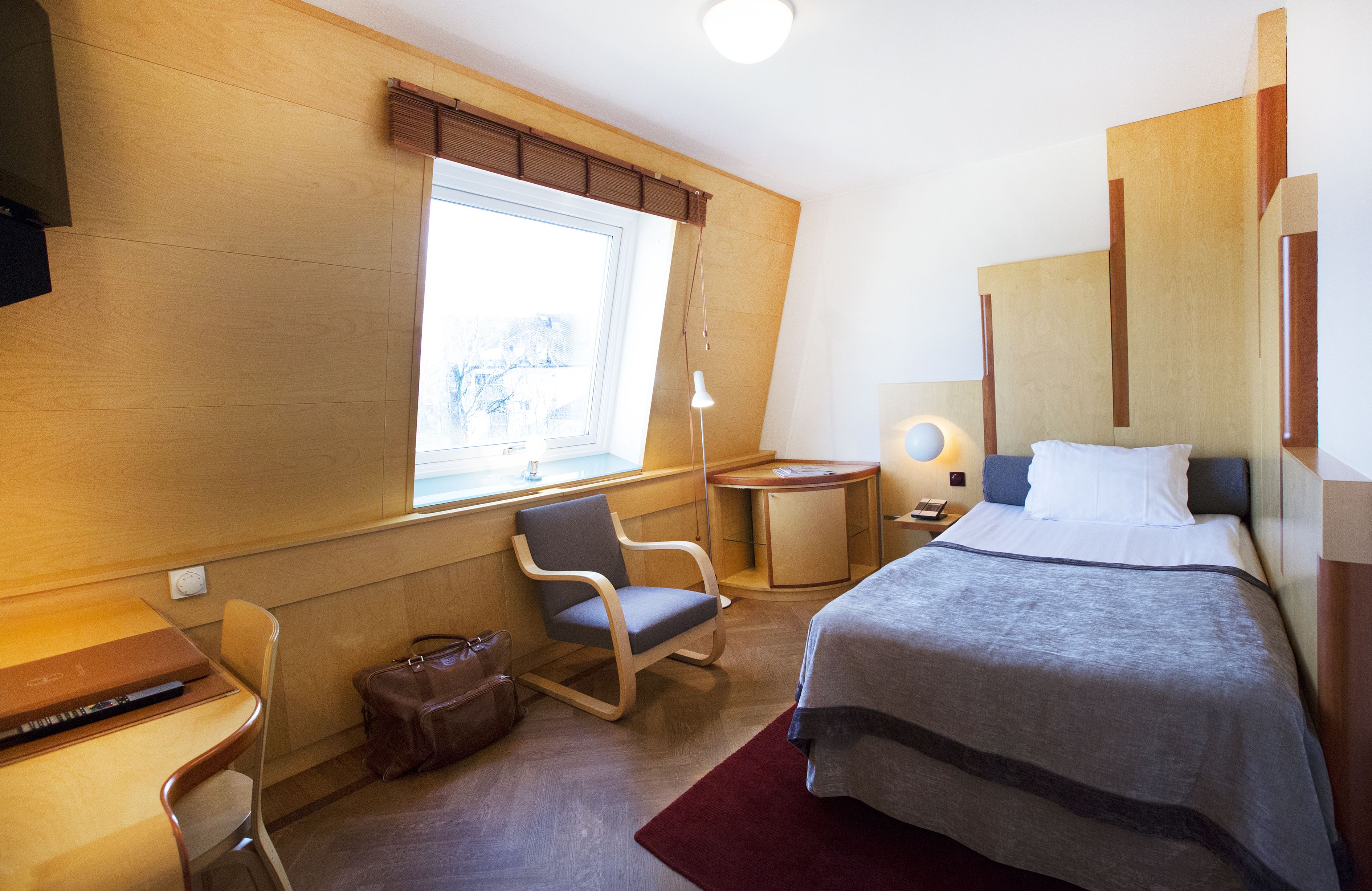 A hotel room with a bed, a desk, a chair, a window, and a lamp.