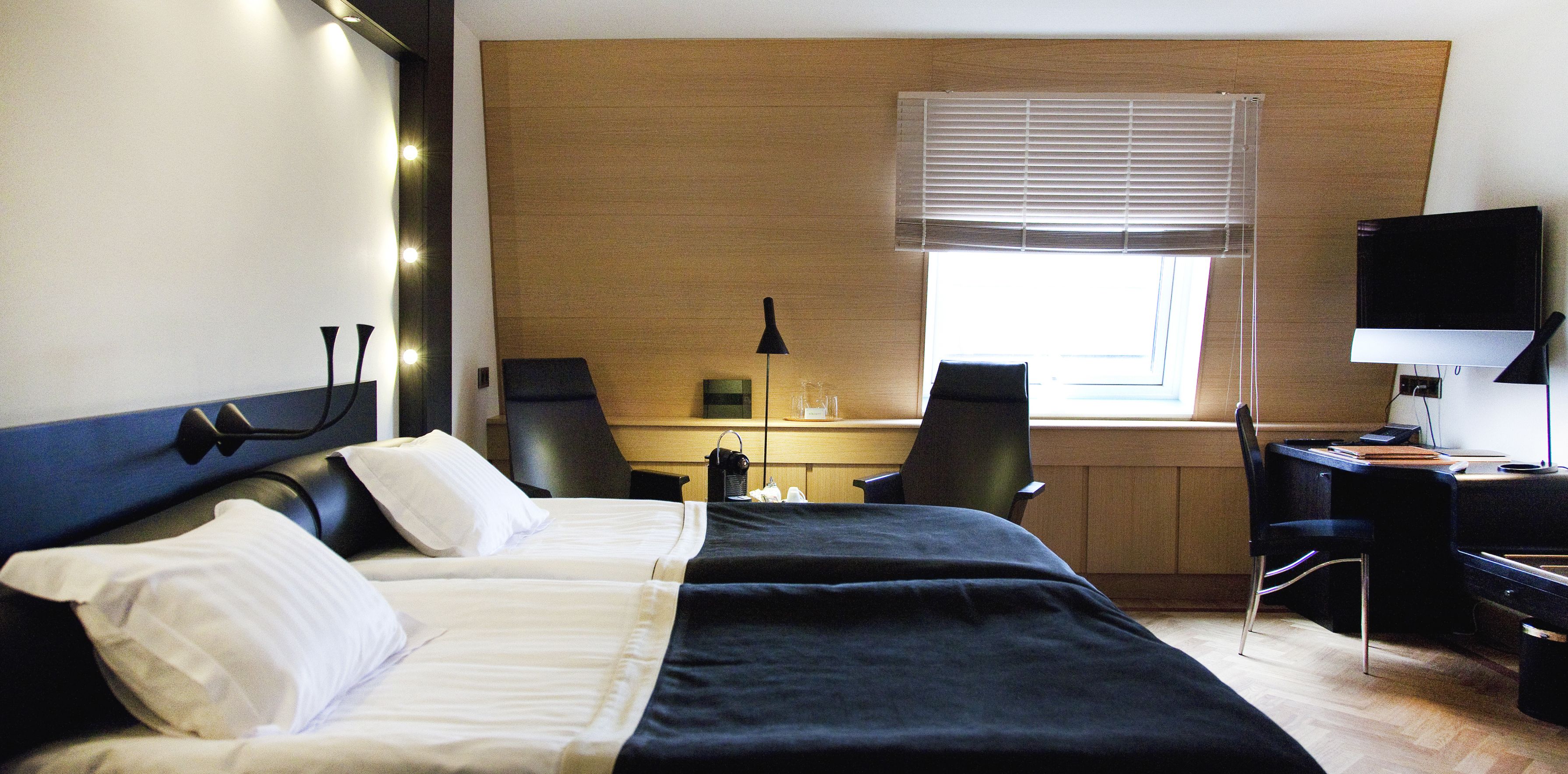 A hotel room with a bed, two chairs, a desk, a TV, and a window with blinds.