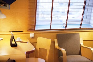 Habitación de hotel con una mesa de madera, dos sillas, una lámpara y un ventanal con persianas.