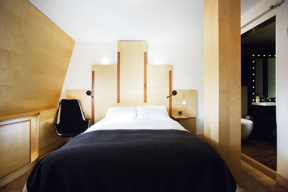 A modern hotel room with a bed, bedside tables, a chair, and a bathroom visible through an open door.