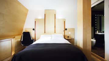 A modern hotel room with a bed, bedside tables, a chair, and a bathroom visible through an open door.