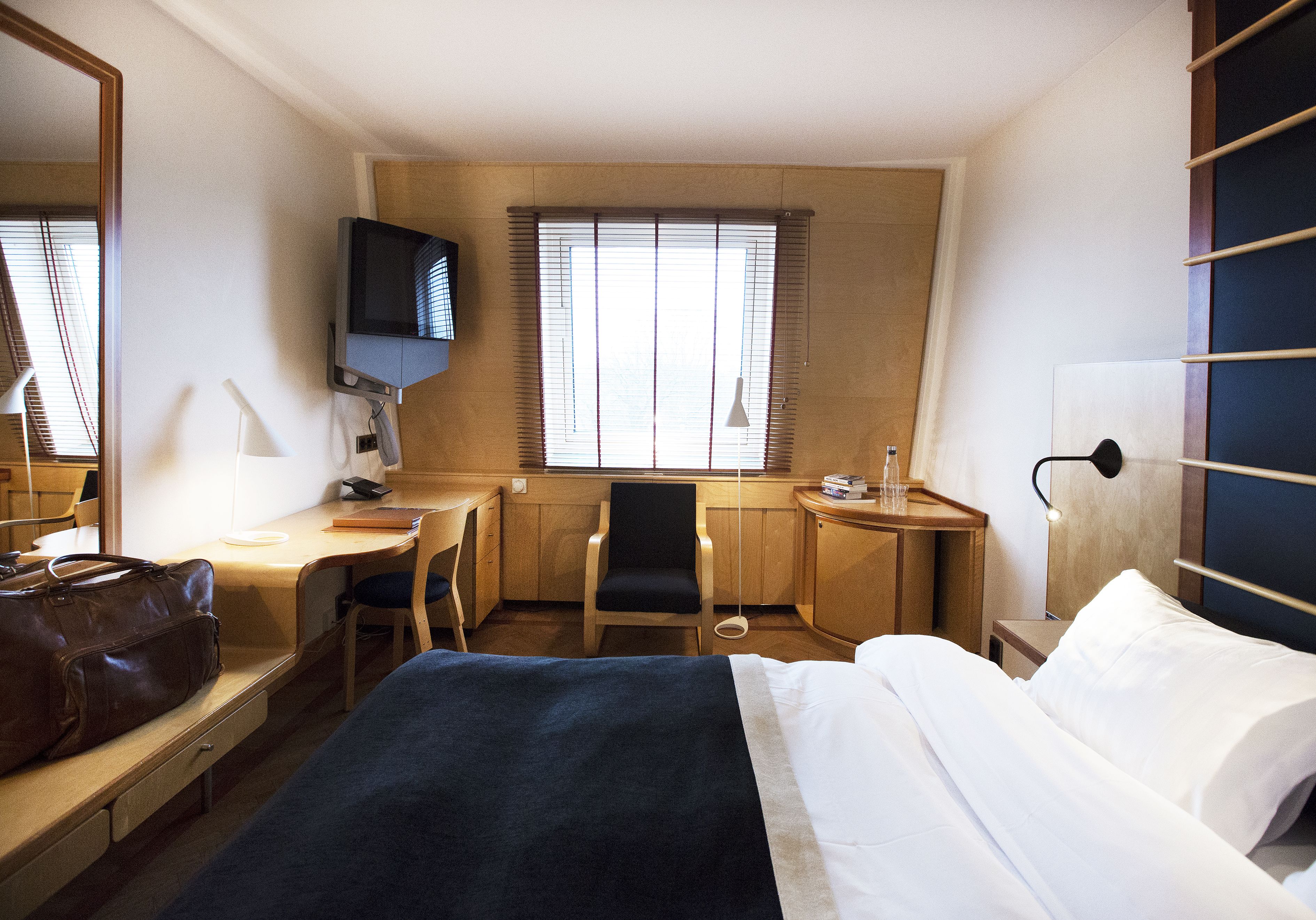 Ein Hotelzimmer mit Bett, Schreibtisch, Stuhl, Fernseher und einem Fenster mit Jalousien.
