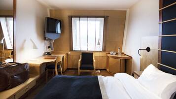 Ein Hotelzimmer mit Bett, Schreibtisch, Stuhl, Fernseher und einem Fenster mit Jalousien.