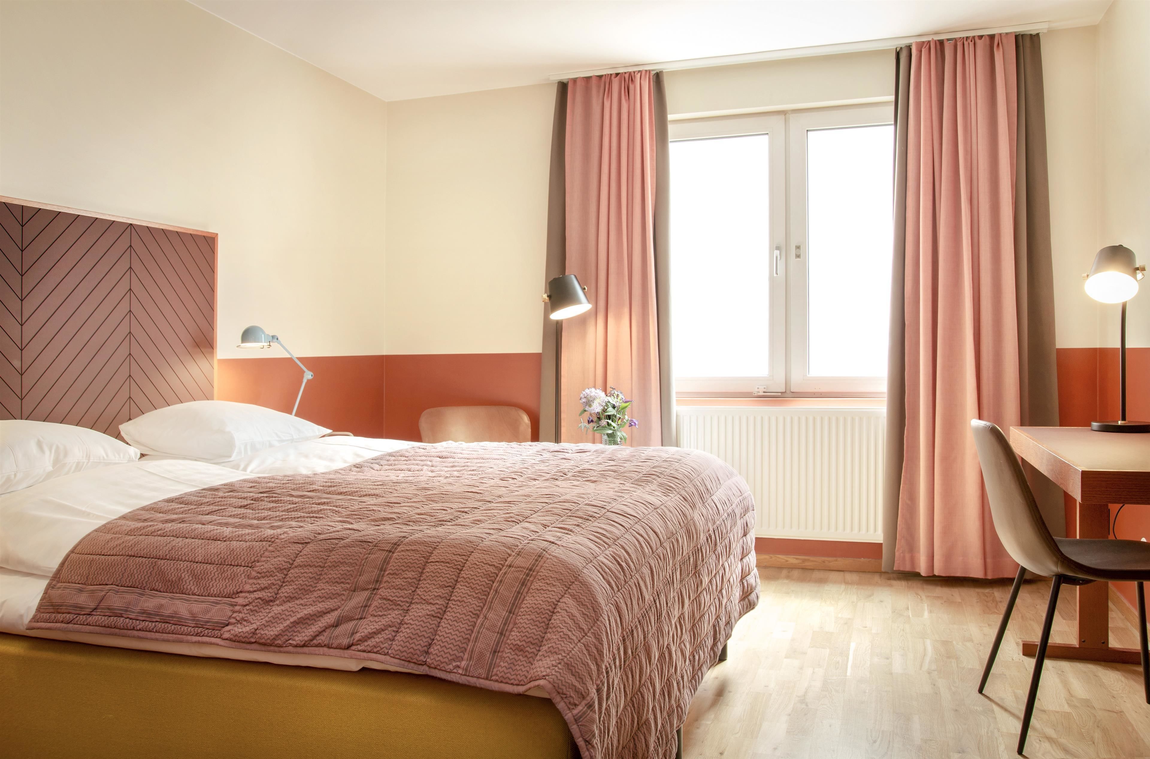 A hotel room with a bed, a desk, a chair, a lamp, and a window with curtains.