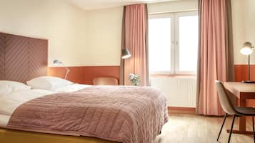 A hotel room with a bed, a desk, a chair, a lamp, and a window with curtains.