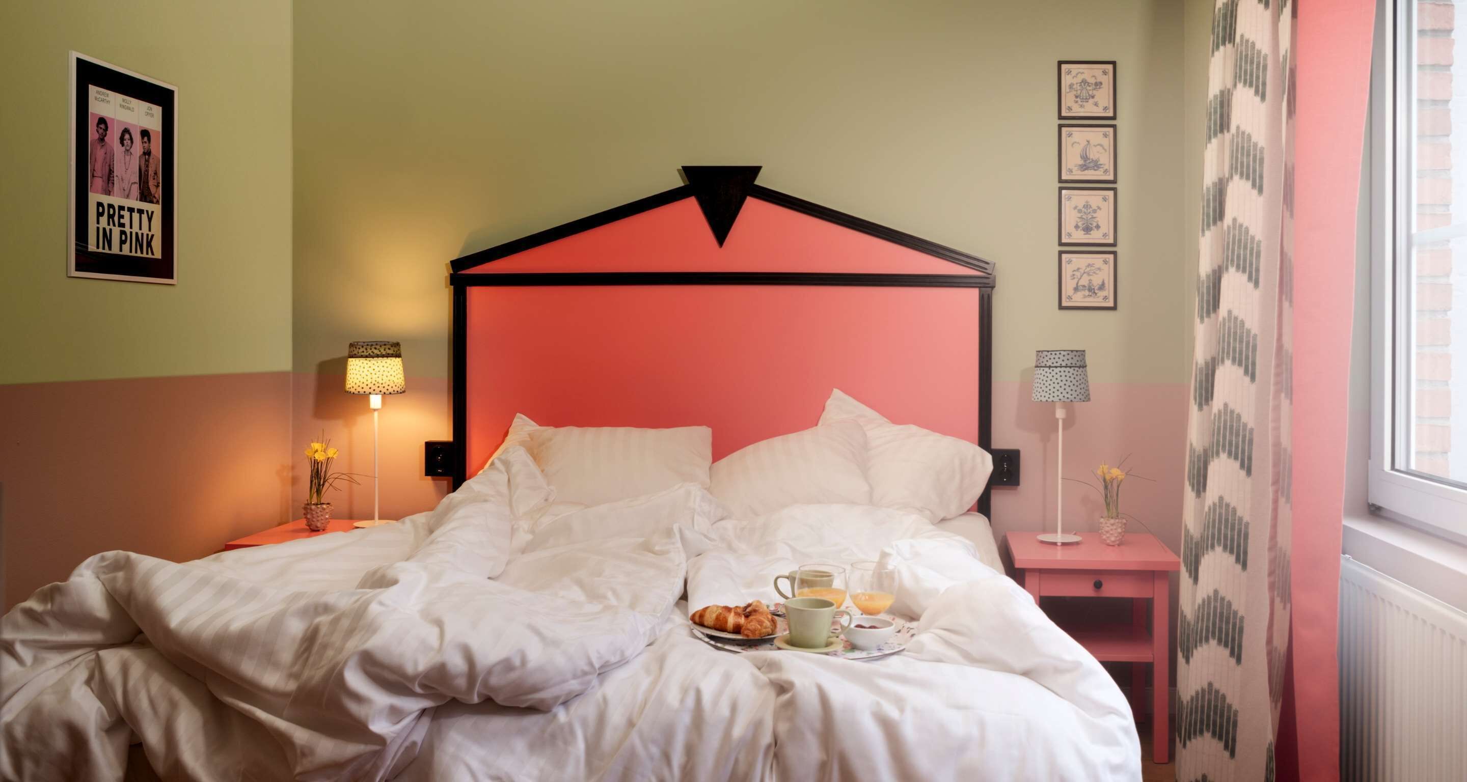 A neatly made bed with white bedding, a pink headboard, and a bedside table with a lamp and a vase of flowers.