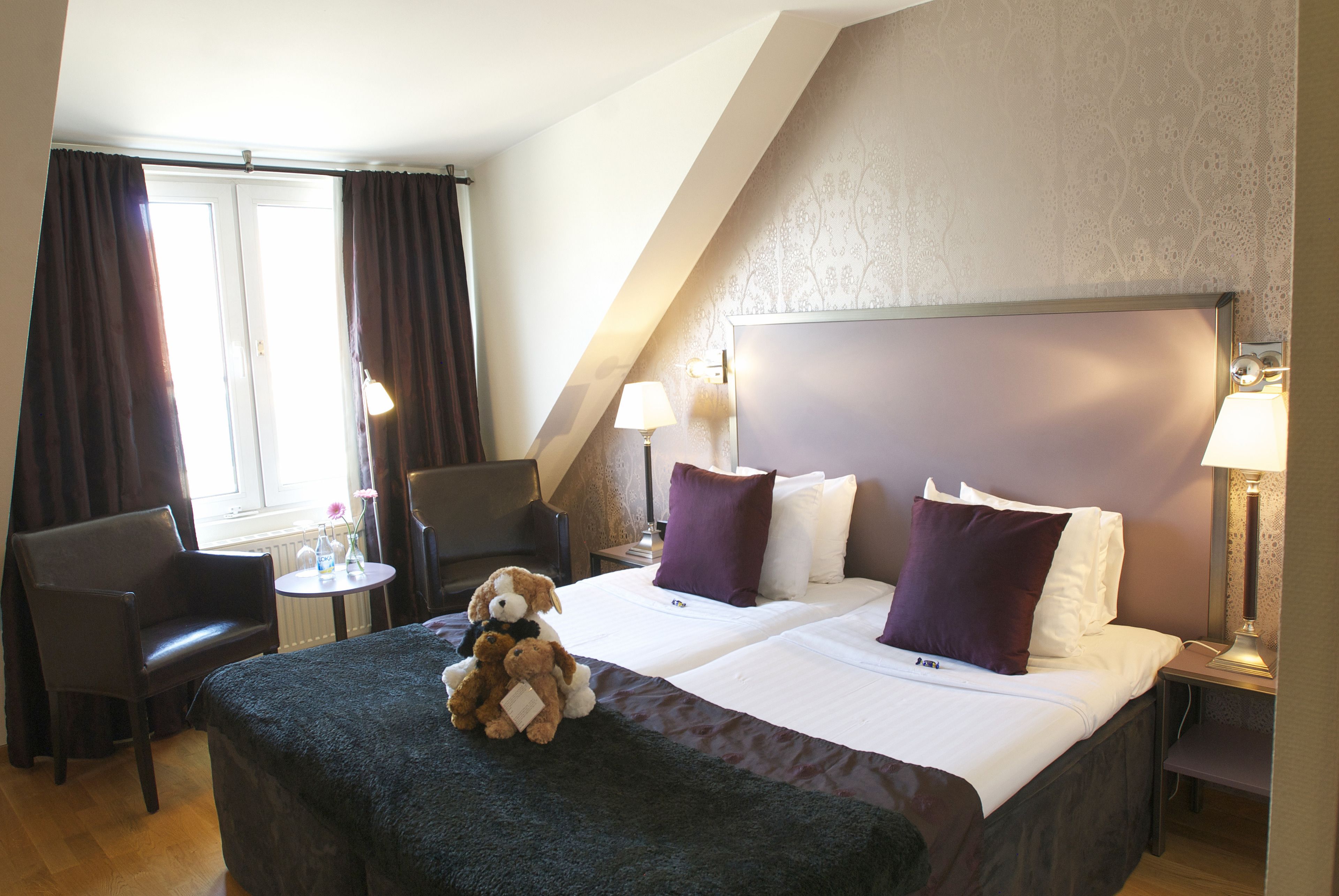 A hotel room with two beds, a sitting area with a chair, a small table, and a lamp.