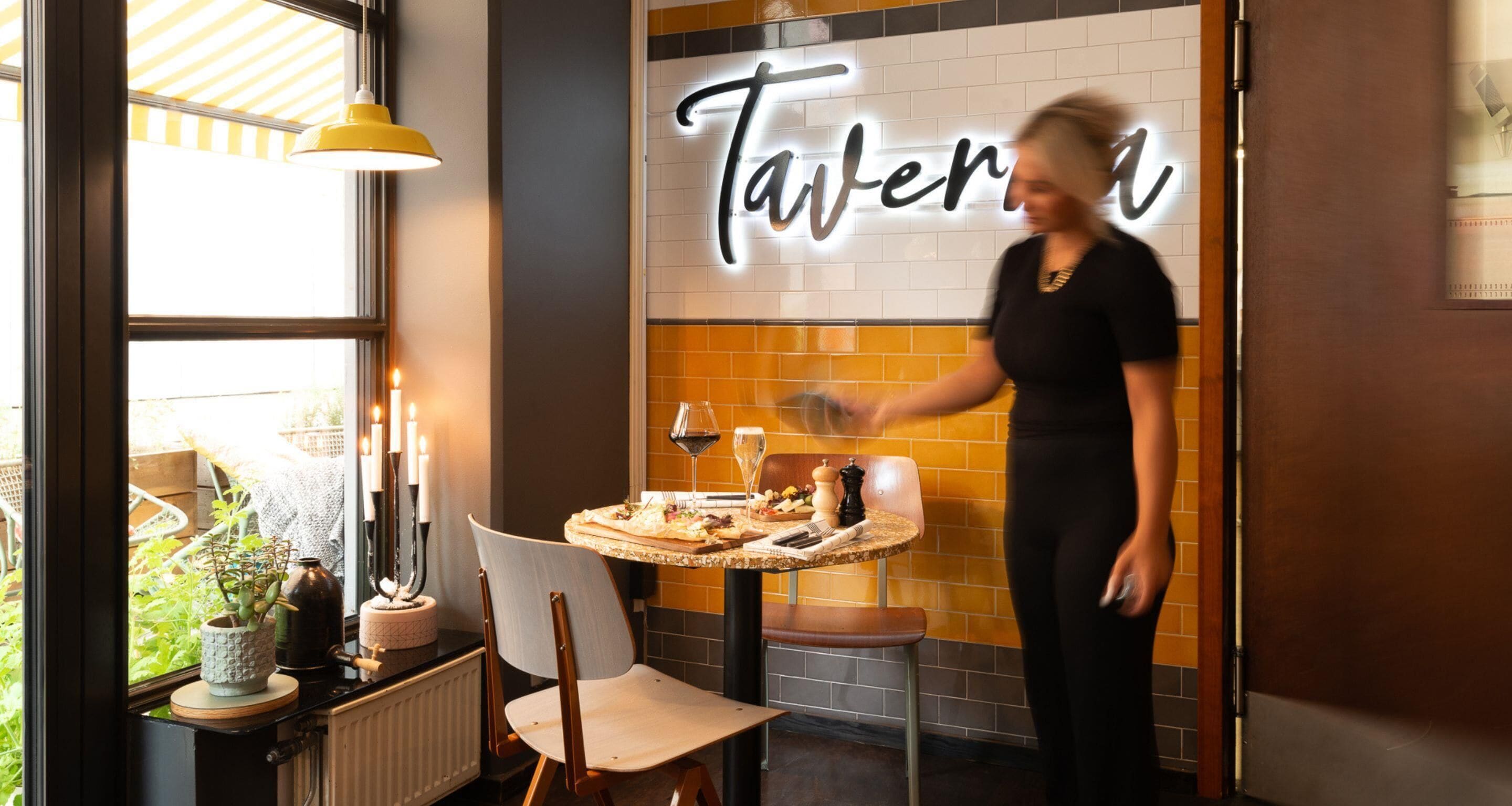 L’intérieur d’un restaurant, avec une enseigne au néon indiquant « Taverna », une table dressée avec de la nourriture et du vin, et une fenêtre donnant sur un paysage verdoyant.