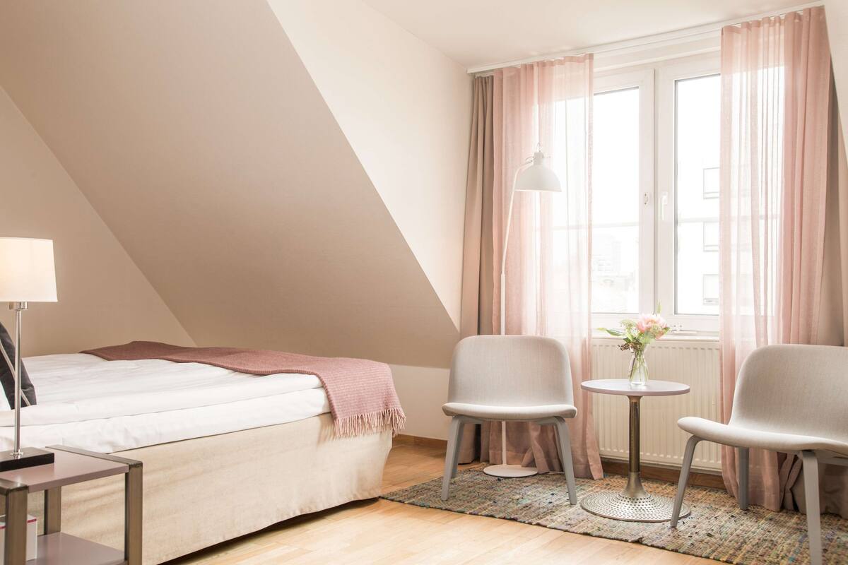 A bedroom with a bed, two chairs, a small table, and a window with curtains.