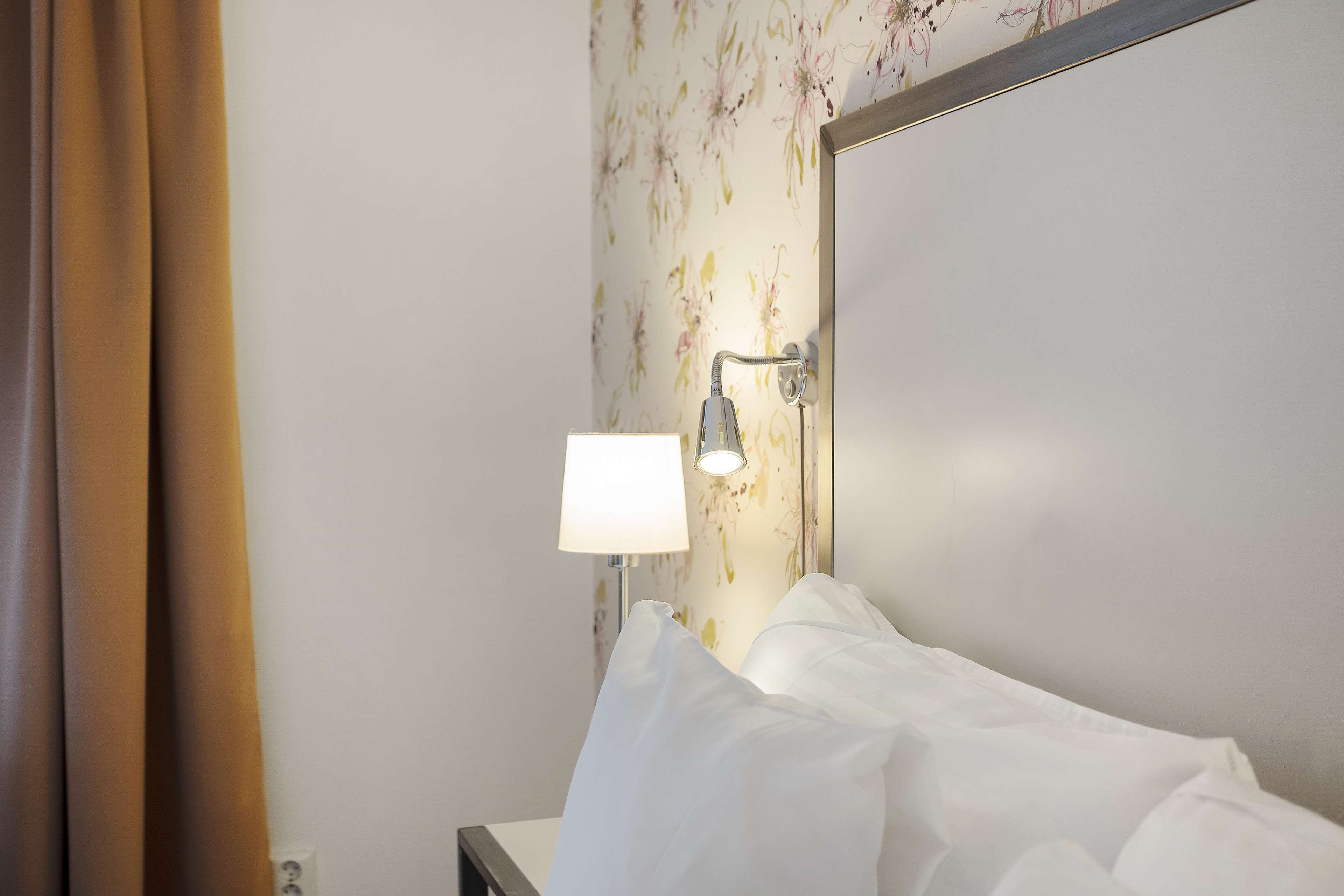 A hotel room with a bed, a nightstand, a lamp, and a wall with floral wallpaper.
