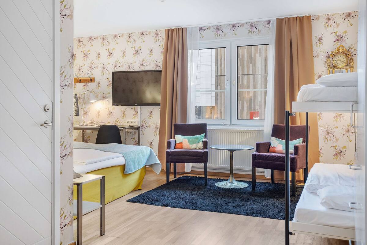 A hotel room with a bed, a desk, two chairs, a small table, a TV, and a window with curtains.