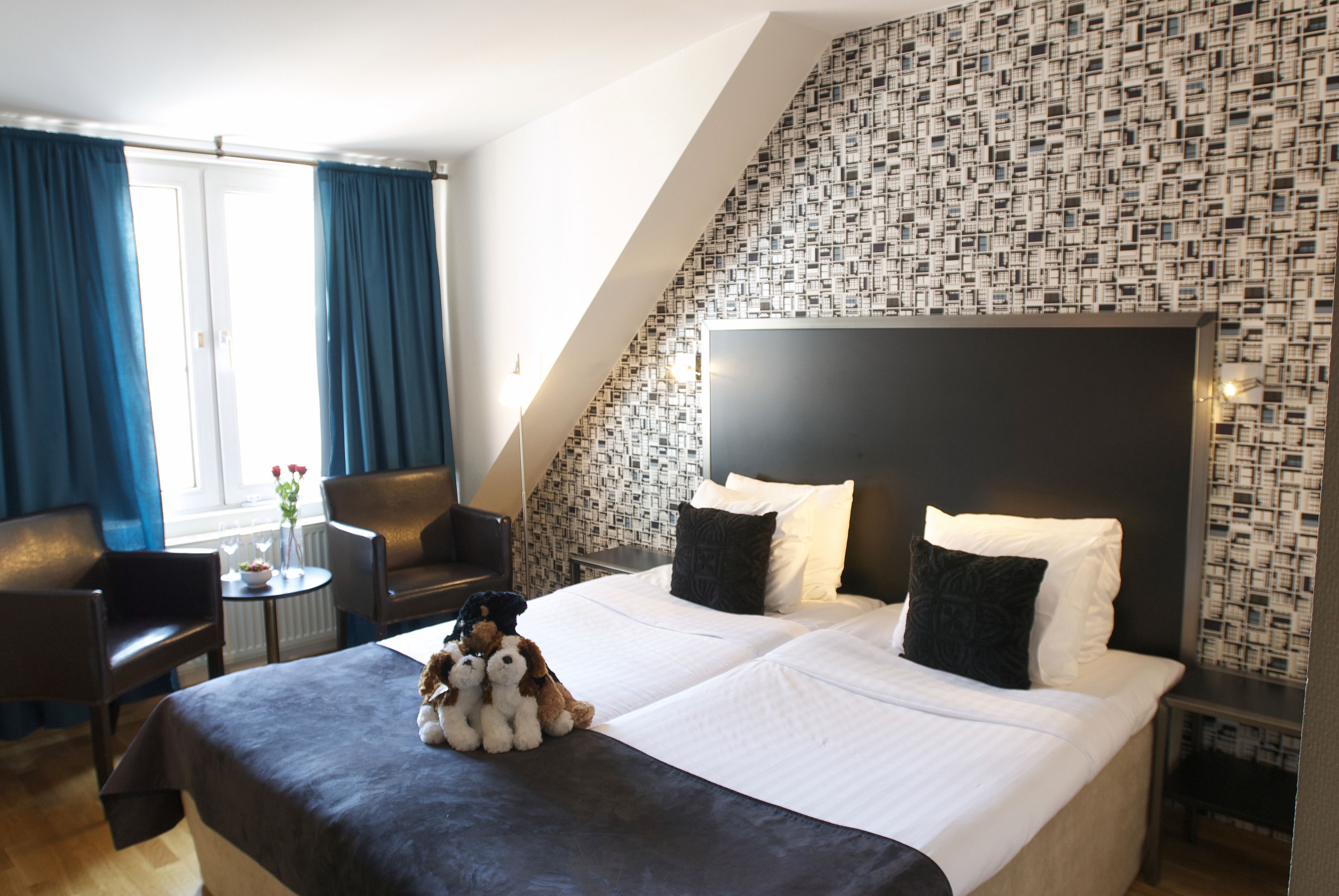 Une chambre d’hôtel avec un grand lit, deux oreillers, une tête de lit, une petite table sur laquelle est posé un vase de fleurs, une chaise et une fenêtre avec des rideaux.