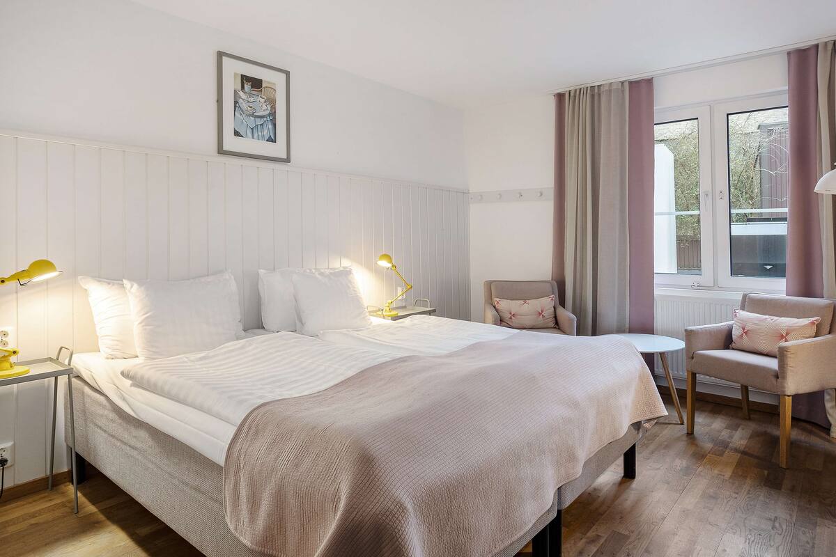 A hotel room with a large bed, bedside tables with lamps, a small round table, a chair, and a window with curtains.