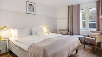 Habitación de hotel con una cama grande, mesitas de noche con lámparas, una pequeña mesa redonda, una silla y un ventanal con cortinas.