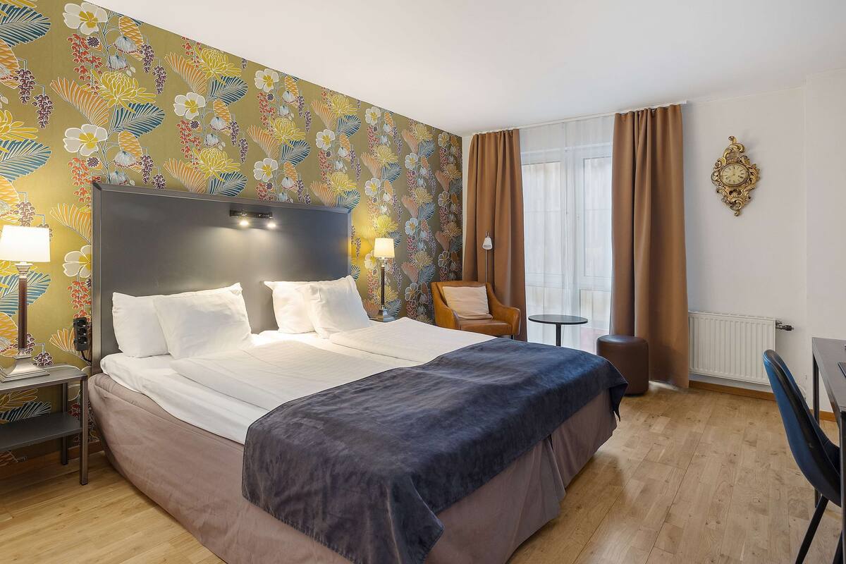 A hotel room with a large bed, a chair, a desk, and a wall clock.
