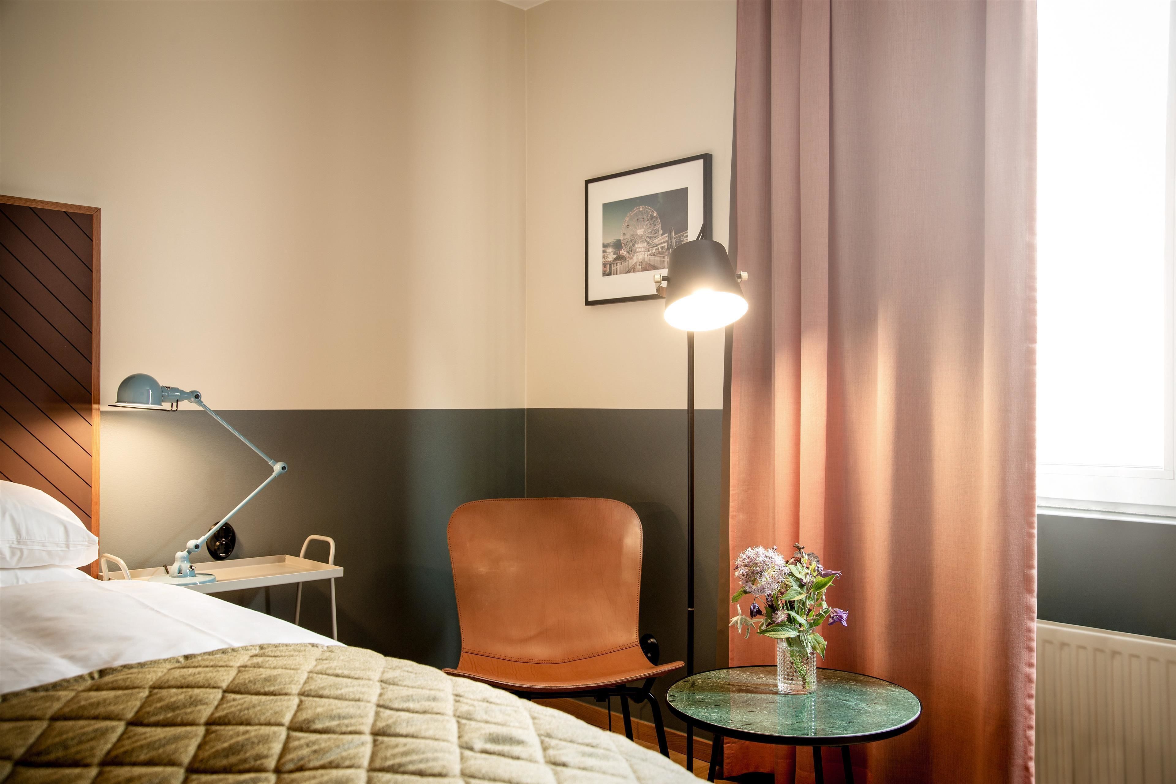 A hotel room with a bed, a desk, a chair, a lamp, a picture on the wall, and a window with curtains.