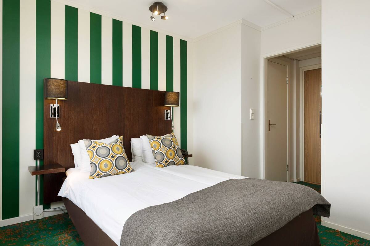 A hotel room with a large bed, green and white striped wall, two bedside lamps, and a wooden door.