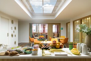 A breakfast buffet with various food items and beverages, a seating area with orange chairs, and a large window with a view of the sky. - ProfilHotels Garden (Malmö)