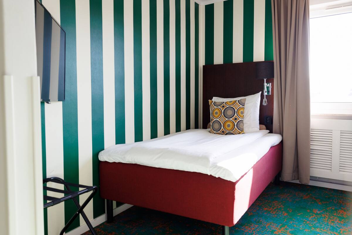 A room with a striped wall, a bed with a red headboard, and a window with curtains.
