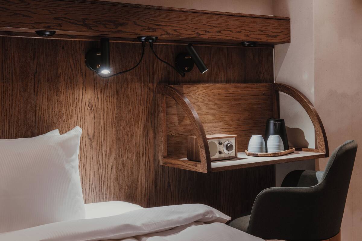 A hotel room with a wooden headboard, a bed with white linens, a small table with a radio and cups, and a black chair.