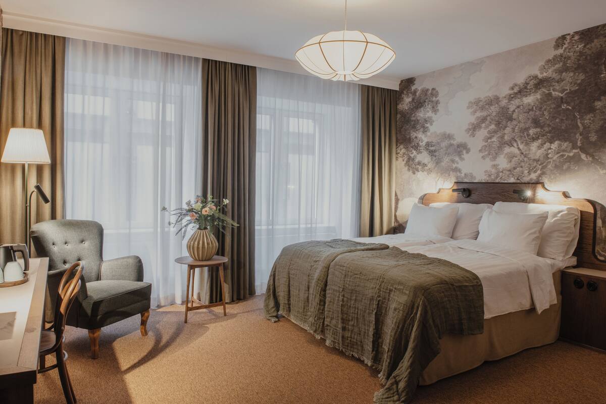 A hotel room with a large bed, a chair, a small table with a vase, and a lamp.