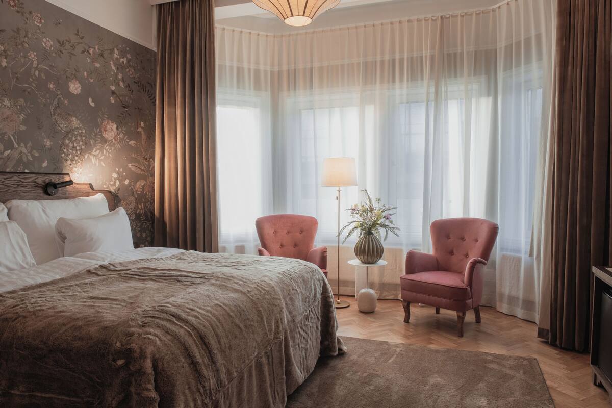 A bedroom with a large bed, two armchairs, a side table with a lamp, and a vase with flowers.