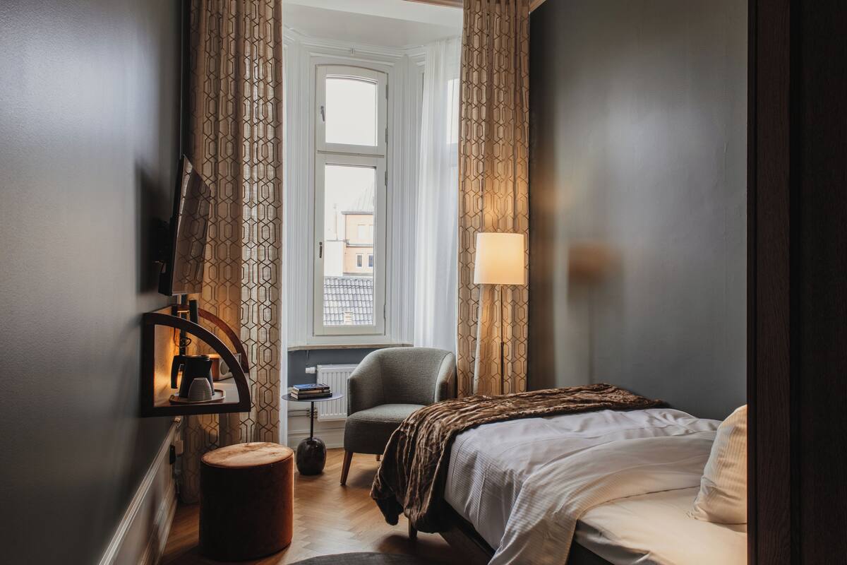 A hotel room with a bed, a chair, a small table with a lamp, and a view of the outside through a window.