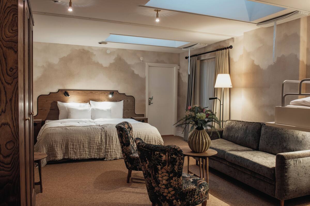 A hotel room with a bed, two chairs, a sofa, a small table, and a lamp. There is a skylight above the bed and a door leading to another room.